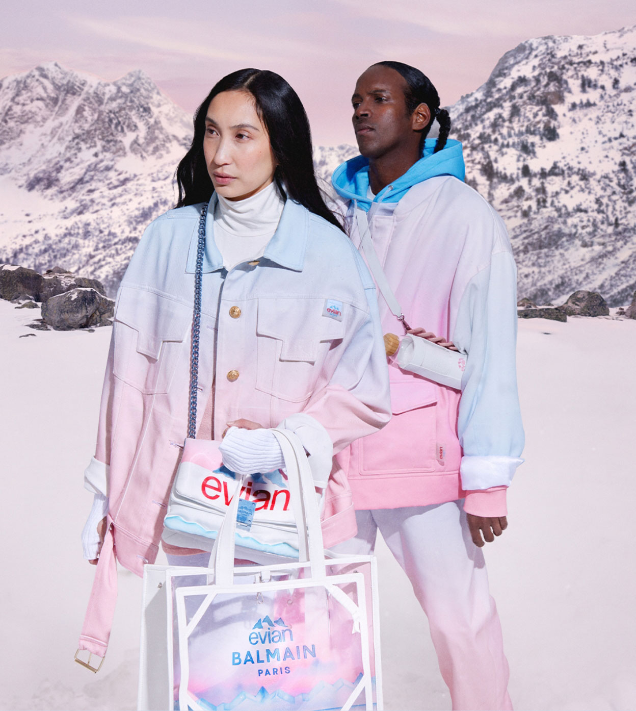 A man and a woman posing in outerwear holding two bags from the evian and Balmain collection.